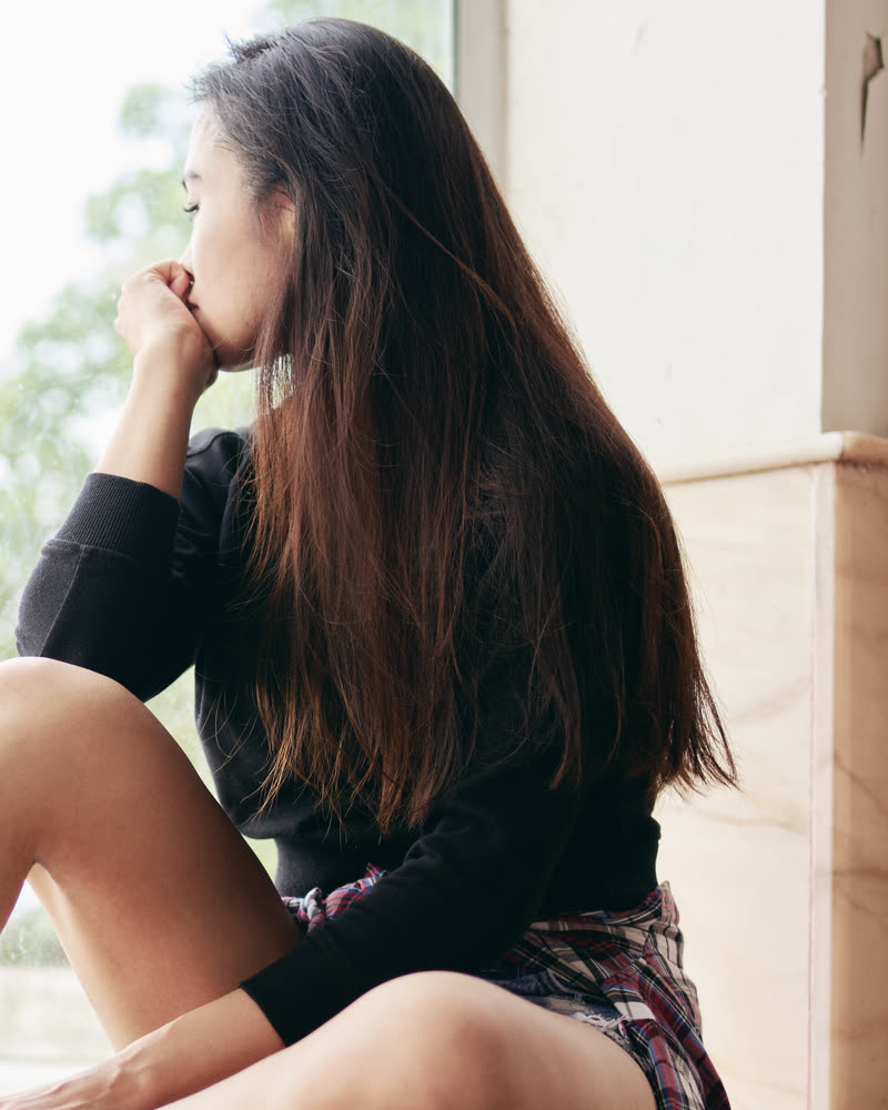 Mujer sentada junto a una ventana, pensativa, con la mano en el mentón